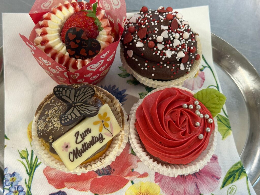 Assorted Valentine-themed cupcakes on a decorative plate: a strawberry-topped cupcake with red-and-white swirled frosting and heart-shaped chocolate, and a chocolate cupcake covered in white and red heart sprinkles.