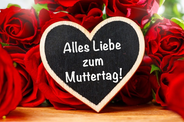 Heart-shaped chalkboard with the German message 'Alles Liebe zum Muttertag!' surrounded by red roses on a wooden surface.