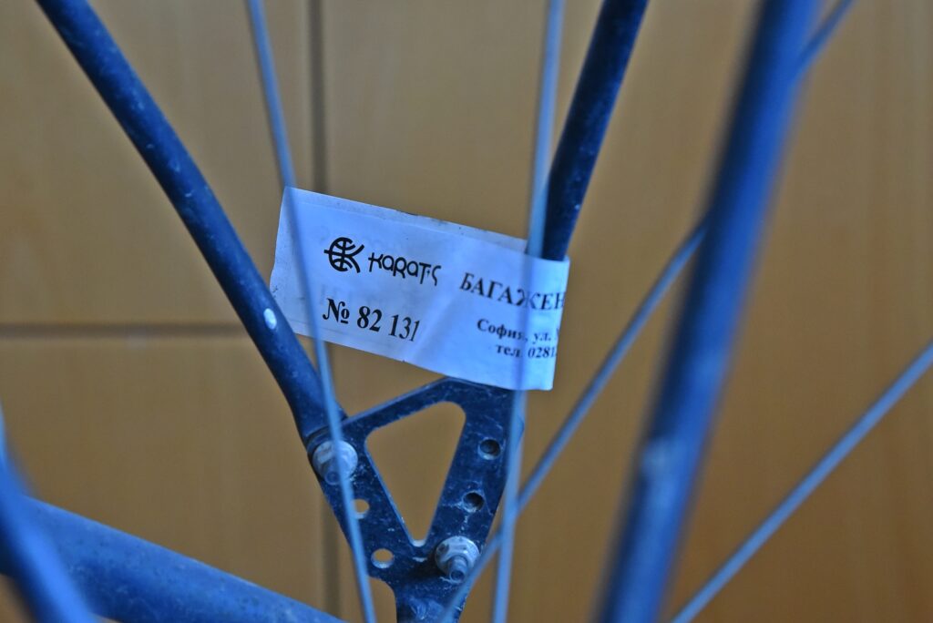 Close-up of a blue bicycle frame with a white tag attached between the tubes, showing Cyrillic text and the number 82 131