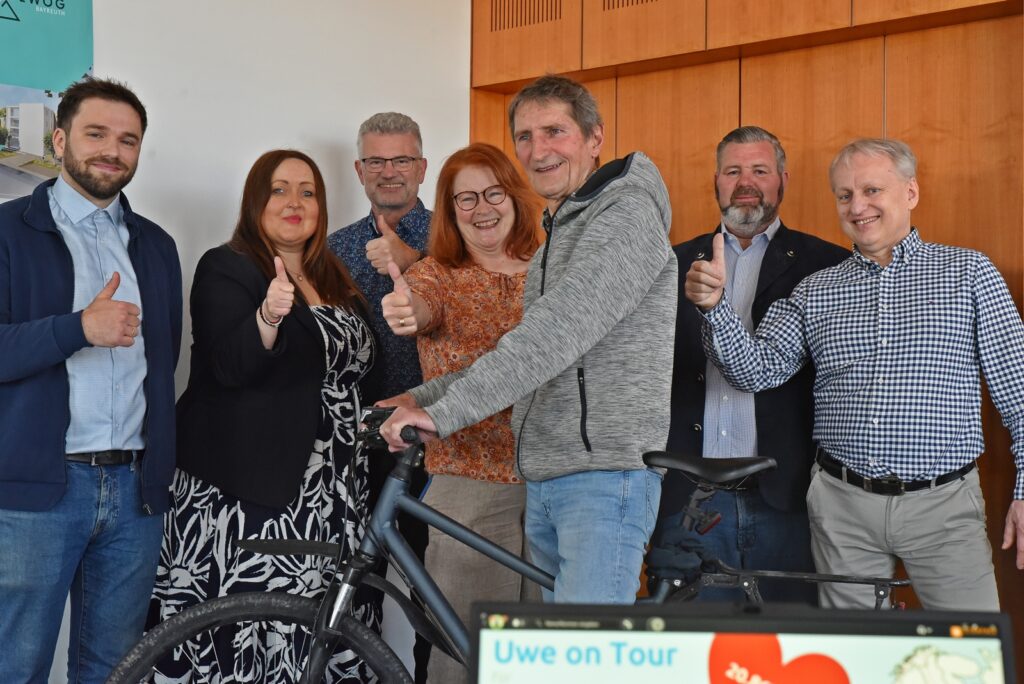 Seven adults indoors, smiling and giving thumbs up around a black bicycle in front of a wood-paneled wall.