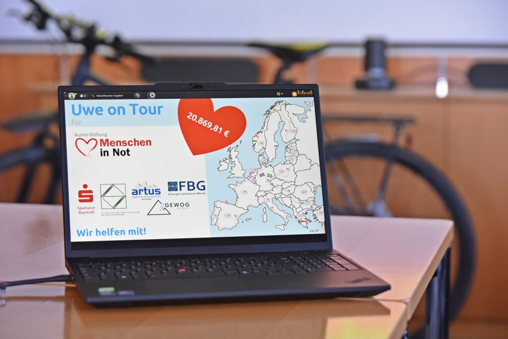 Laptop displaying a Europe map and a large red heart graphic with 'Uwe on Tour' title and logos on a desk.
