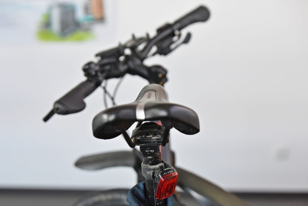 Rear view of a bicycle saddle and seat post with handlebars blurred in the background; a red rear light is visible behind the seat.