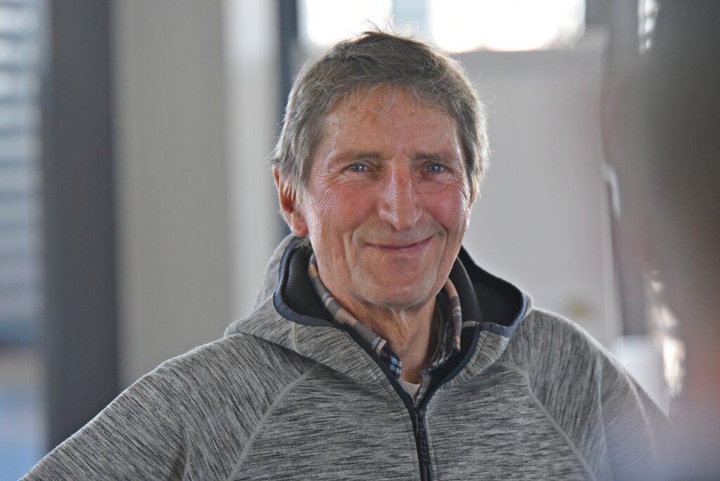 Portrait of a smiling older man with short gray hair wearing a gray zip-up hoodie indoors.